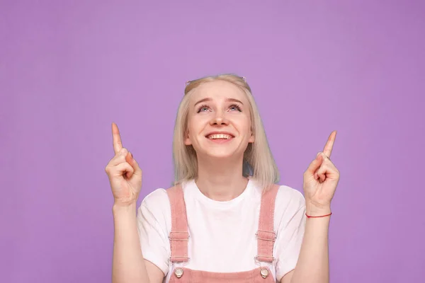 Close-up portret van een vrolijk tienermeisje met licht haar kijkt op een paarse achtergrond en toont een vinger. Gelukkig blond toont vingers omhoog in een lege ruimte en glimlacht, geïsoleerd. — Stockfoto