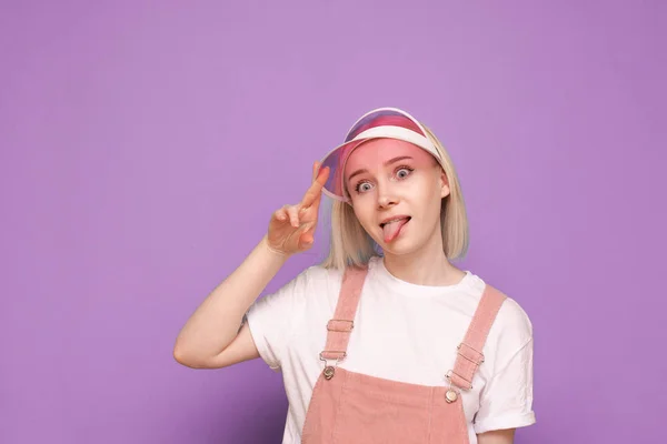 Portrét legrační dívky v pěkných neformálních šatech, s růžovou čepicí a legračním obličejem na purpurovém pozadí. Vtipné teen dívka pózuje na kameru, izolované detailní portrét. — Stock fotografie