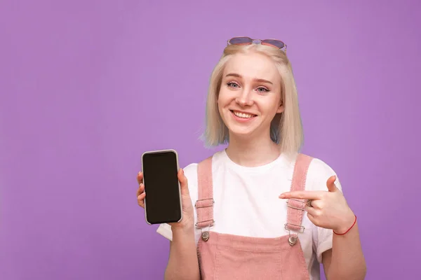 Joyeux blond fille tient un smartphone avec un écran noir, montre un doigt sur l'écran et regarde dans la caméra. sourire adolescent fille montre un doigt sur un smartphone dans sa main — Photo