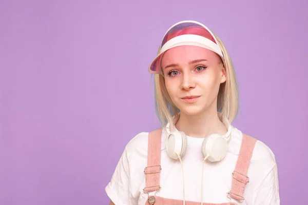 Bella ragazza con le cuffie e un berretto rosa è su uno sfondo blu e guarda la fotocamera, un ritratto da vicino. Ritratto di un'adolescente che posa sullo sfondo di un muro viola. Isolato . — Foto Stock