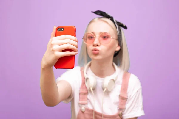 Pembe gözlüklü komik bir kızın yakın plan portresi mor arka planda komik bir suratla akıllı bir telefondan selfie çekiyor. Akıllı telefona odaklan.. — Stok fotoğraf
