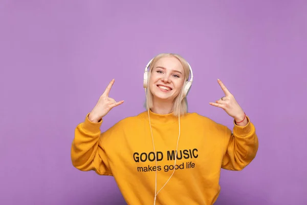 Щаслива блондинка в помаранчевій сорочці і навушники на фіолетовому фоні, слухає музику і показує знак важкого металу, дивиться в камеру і посміхається. Ізольовані — стокове фото