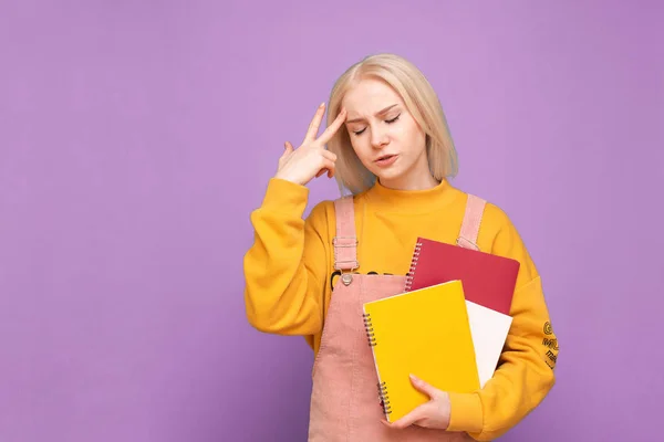 疲惫的女学生，手里拿着书本和笔记本，双目紧闭地站在紫色的背景上。 头痛的学生被隔离在粉红的背景下. 学习概念. — 图库照片