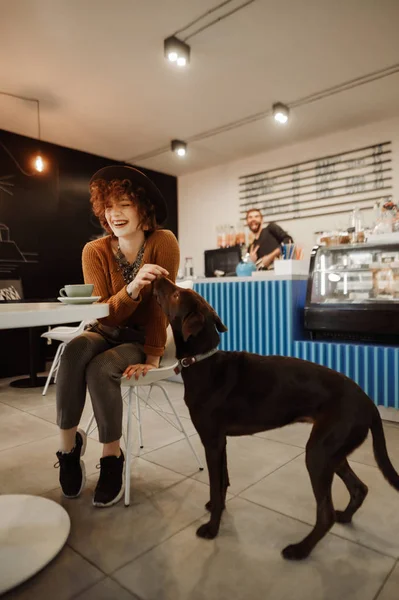 Πορτρέτο του χαρούμενος κορίτσι κάθεται στο τραπέζι σε ζεστό καφέ με φλιτζάνι καφέ και παίζει με το σκυλί, χαμογελώντας και κοιτάζοντας μακριά. Hipster κορίτσι με κομψά ρούχα ξοδεύει χρόνο στο φιλικό προς τα ζώα καφέ. — Φωτογραφία Αρχείου