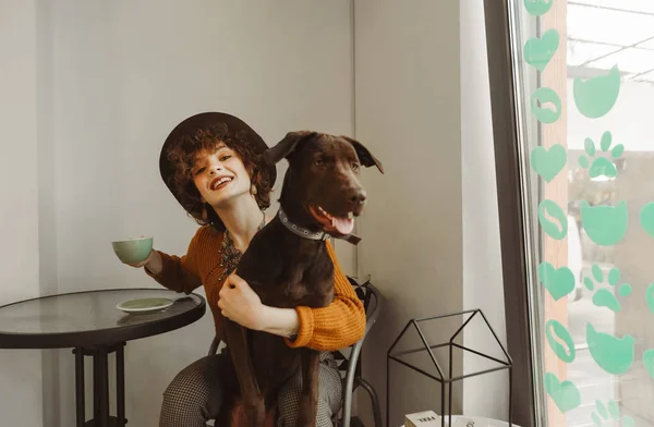 En glad pige i en hat sidder på en cafe med en kop kaffe og en hund på skødet, kigger ind i kameraet og smiler. Ejeren med et kæledyr i hænderne spiser morgenmad på cafeen . - Stock-foto