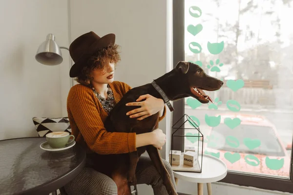 Retrato de chica hipster en la cafetería con taza de café en la mesa abrazando perro. Señora con ropa elegante jugando con un perro en una acogedora cafetería junto a la ventana. Café es amigable con animales —  Fotos de Stock