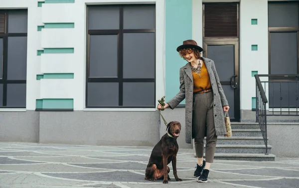 Leende snygg kvinna och brun hund i koppel stå på bakgrunden av en byggnad, flicka ler och tittar på ett husdjur.Fashionabla porträtt av en lycklig dam i en hatt på en promenad med valpar i staden — Stockfoto