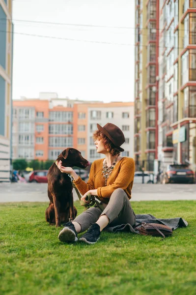 Élégante fille hipster assis sur la pelouse avec un chien sur un fond de bâtiments modernes et joue avec un animal de compagnie. Portrait vertical d'un propriétaire d'animal marchant sur une promenade, assis sur une pelouse et relaxant . — Photo