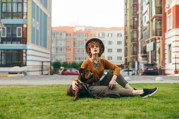 stock image Portrait attractive girl sitting with a puppy in the yard on the lawn, looking into the camera, wearing stylish clothes and a hat. Hipster girl relaxing with dog on grass,modern building background