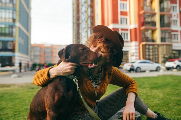 Cute girl w kapeluszu siedzi na trawniku z brązowym psem, przytulanie i całowanie. Kocham właściciela za psa. Kobieta w kapeluszu przytula psa siedzącego na trawie na podwórku. Miłość do zwierząt domowych. — Zdjęcie stockowe
