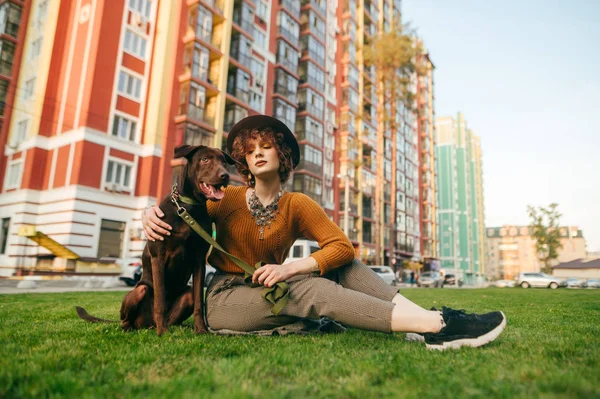 Attraktive Hipster-Mädchen mit Hut und stilvolle Kleidung sitzt auf dem Rasen im Hof und umarmt einen Welpen. niedliche Dame spielt mit braunen Welpen auf Gras auf Gebäuden Hintergrund. Haustierkonzept. — Stockfoto