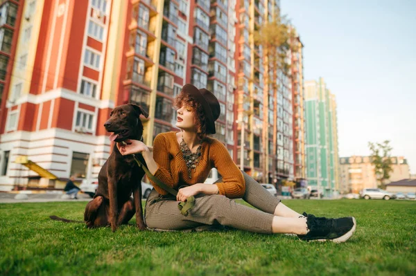 Chica sonriente en ropa elegante jugando con un lindo perro marrón en el césped en el patio sobre un fondo de edificios de colores modernos. La dama de sombrero se sienta en la hierba y sujeta al perro con correa. Ocio con mascota . —  Fotos de Stock