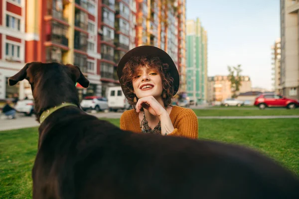 Portret szczęśliwej dziewczyny z psem na trawniku na podwórku na tle pejzażu miejskiego, patrzącej w kamerę i uśmiechniętej. Uśmiechnięta pani ze szczeniakiem na spacerze, pozująca przed kamerą — Zdjęcie stockowe