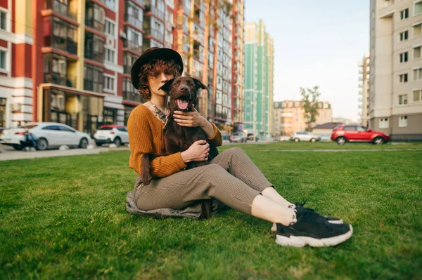 スタイリッシュな女の子は居心地の良い庭で芝生の上に座って、犬を抱き、カメラを見ています。草の上でリラックスした帽子の女性と彼女の腕の子犬と都市の背景にあります。ペットと一緒に歩く. — ストック写真