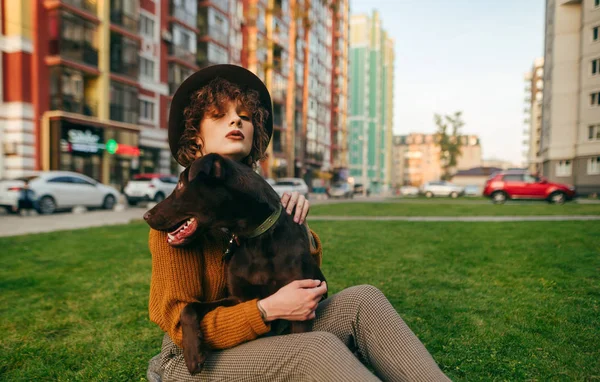 A senhora atraente no chapéu senta-se no quintal no gramado com o cão em suas mãos e posa na câmera no fundo da paisagem urbana com grama e edifícios. Hipster menina abraça um cão em uma caminhada e olha para a câmera — Fotografia de Stock