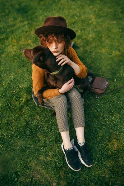 Porträt eines Mädchens, das auf einem grünen Rasen sitzt und einen schönen braunen Hund umarmt, Draufsicht. attraktive Dame umarmt und streichelt den Welpen. Haustiere. vertikales Foto. — Stockfoto