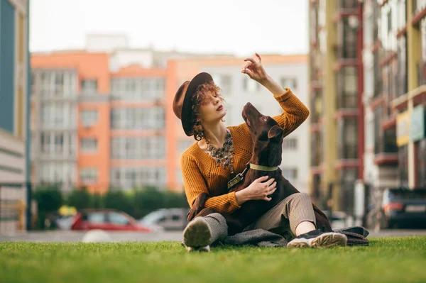 Atrakcyjna stylowa hipsterka w kapeluszu bawiąca się psem siedzącym na trawniku na rozmytym tle z budynkami, szczeniak patrzący na paszę w ręku właściciela. Wypoczynek z psem na spacerze po podwórku. — Zdjęcie stockowe