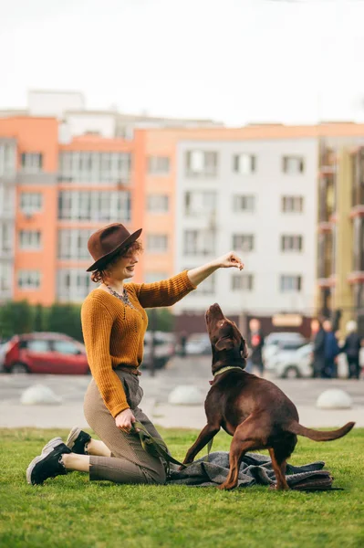 Elegante chica hipster viste al perro en el césped contra el bac —  Fotos de Stock