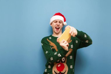 Noel Baba şapkası ve yeşil Noel süveteri giyen etkileyici genç adam mavi arka planda video oyunları oynuyor, akıllı telefon ekranına bakıyor ve bağırıyor. Duygusal genç oyuncu gezici oyunlar ve Noel oynuyor