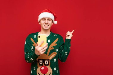 Gülümseyen genç bir adam elinde bir akıllı telefonla kırmızı arka planda duruyor, kameraya bakıyor, gülümsüyor ve fotokopi alanını işaret ediyor, Noel süveteri ve Noel Baba şapkası giyiyor. İzole edilmiş