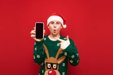 Noel Baba şapkalı ve Noel Baba kazaklı adam elini akıllı telefon ekranında gösteriyor ve kırmızı arka plandaki izole kameraya bakıyor. Şok olmuş adam kameraya siyah akıllı telefon ekranı gösteriyor..