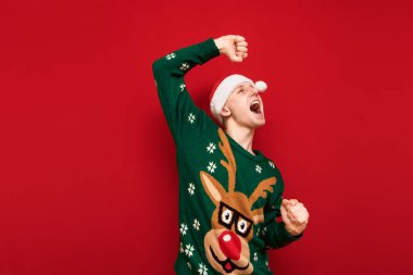 Noel Baba şapkası ve yeşil kalın süveter giyen neşeli genç adamın fotoğrafı. Eğleniyor ve kırmızı arka planda, başka tarafa bakıyor ve gülümsüyor. Şenlik elbiseli adam dans ediyor. Noel partisi.