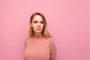 Pembe kazaklı güzel sarışın kız pembe bir arka planda izole edilmiş, boş bir yere bakıyor ve gülümsüyor. Pozitif kadın renkli arka plandan başka tarafa bakıyor. Kopyalama alanı