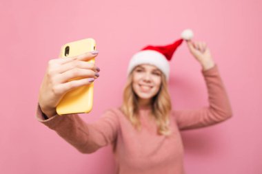 Sarı kapaklı akıllı telefona odaklan. Noel şapkalı sevimli sarışın kız pembe arka plandaki akıllı telefondan selfie çekiyor, kameraya bakıyor ve gülümsüyor.