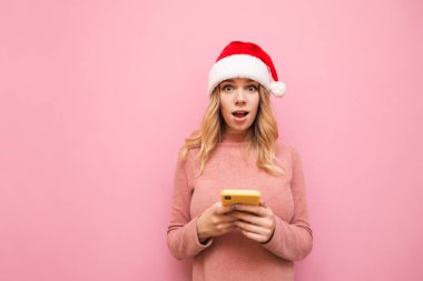 Şok olmuş Noel şapkalı kız akıllı telefon tutuyor ve pembe arka plandaki kameraya şaşırmış görünüyor. Pembe kazaklı, akıllı telefon kullanan, izole edilmiş güzel bir kız. Xmas kavramı