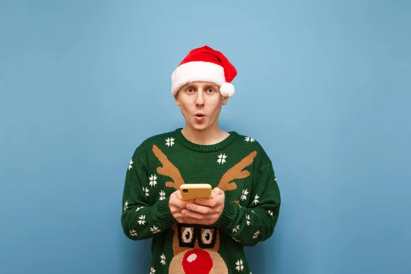 Uomo sorpreso in maglione di Natale e cappello Babbo Natale stand con smartphone in mano su sfondo blu, fai da te in macchina fotografica. Divertente ragazzo in abiti di Natale utilizza smartphone su sfondo blu . — Foto Stock