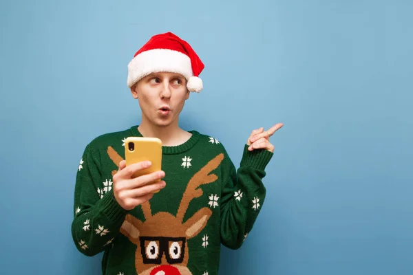 Sorpreso giovane uomo in cappello di Babbo Natale e caldo maglione stand con smartphone in mano sullo sfondo blu, guarda lontano lo spazio vuoto e punti con il dito. Isolato. Copia spazio — Foto Stock