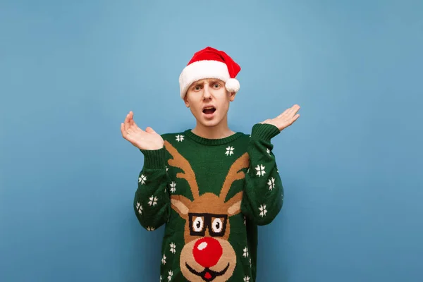 Confused young man in a Christmas hat and sweater isolated on a blue background, looks at the camera in surprise and spreads his hands to the sides. Studio portrait of confused guy in Christmas. — Stock Photo, Image