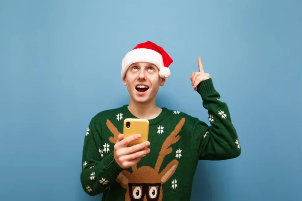 Cheerful young man in christmas sweater and hat isolated on blue background with smartphone in hand, looks up, smiles and shows finger on copy space. Joyful Guy with phone pointing finger empty space. — Stock Photo, Image