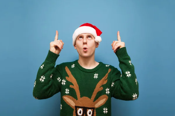 Chockad ung man klädd i en varm jultröja och hatt Santa står mot en blå bakgrund, tittar upp och pekar fingrarna på ett tomt utrymme. Förvånad julkille pekar finger åt snuten — Stockfoto