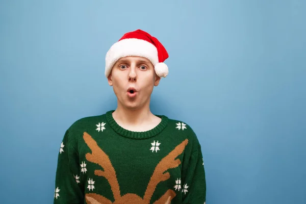 Ritratto ravvicinato di giovane scioccato in cappello di Babbo Natale e maglione di Natale verde si erge su sfondo blu e guarda sorpreso dalla fotocamera con faccia sorpresa. Isolato . — Foto Stock