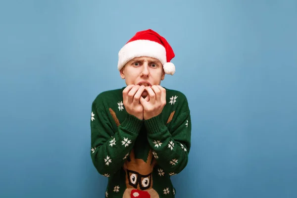 An emotional young man in a hat of Santa and in a sweaty sweater is worried, looking frightened. Closeup portrait of a frightened guy in Christmas, looking shockingly into the camera. Isolated.