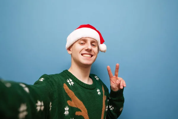 Sonriente joven con sombrero de santa y suéter verde hace selfie de Navidad sobre fondo azul, muestra a la cámara un gesto de paz. Guapo feliz chico haciendo Navidad selfie, aislado . —  Fotos de Stock