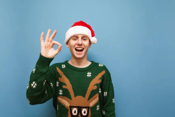 Chico alegre en jersey de navidad verde y sombrero de navidad se encuentra en el fondo azul, muestra gesto OK, mira a la cámara y sonríe. Joven alegre mostrando los pulgares hacia arriba gesto OK —  Fotos de Stock