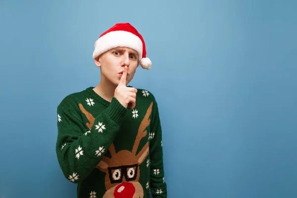 En seriös ung man i julkläder tittar in i kameran och visar en gest av tystnad. Fokuserad kille sätter fingret på läpparna och med ett seriöst ansikte tittar in i kameran, ber han om tystnad — Stockfoto