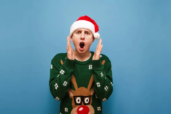 크리스마스 옷을 입고 파란 배경에 충격을 받은 남자의 사진. 미친 Lyceum 이 카메라를 보고 소리를 지르며 산타 모자를 쓰고 녹색의 따뜻 한 스웨터를 입고 있다. 고립됨... — 스톡 사진