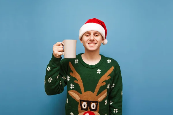 Retrato de joven sonriente en jersey verde cálido y sombrero de santa claus, de pie sobre fondo azul con copa de bebida caliente en la mano, mirando a la cámara y sonriente. Año nuevo y concepto de Navidad —  Fotos de Stock