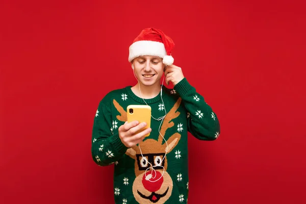 Όμορφος νεαρός άνδρας στέκεται σε ένα κόκκινο φόντο με ένα smartphone στο χέρι του, ακούει μουσική στα ακουστικά και χρησιμοποιεί το Διαδίκτυο, κοιτάζει την οθόνη και χαμογελά. Απομονωμένοι. Χριστουγεννιάτικη ιδέα — Φωτογραφία Αρχείου
