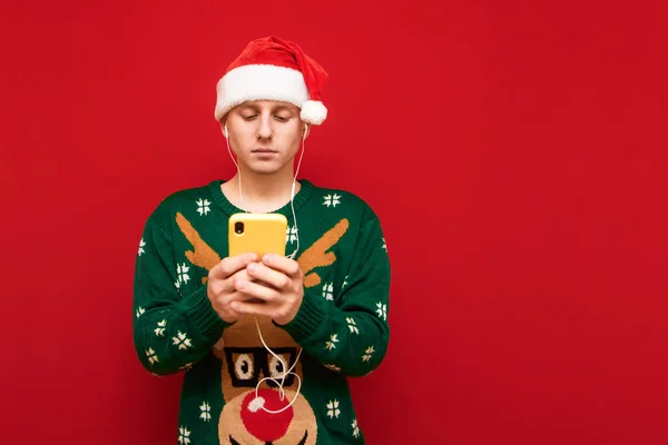 산타 모자와 녹색 크리스마스 옷을 입은 젊은 남성 이 빨간 배경에 서 있고, 진지하게 보이는 스마트폰을 사용하고 헤드폰을 쓰고 음악을 듣고, 산타 모자를 쓰고 녹색의 따뜻 한 스웨터를 입고 있다. — 스톡 사진