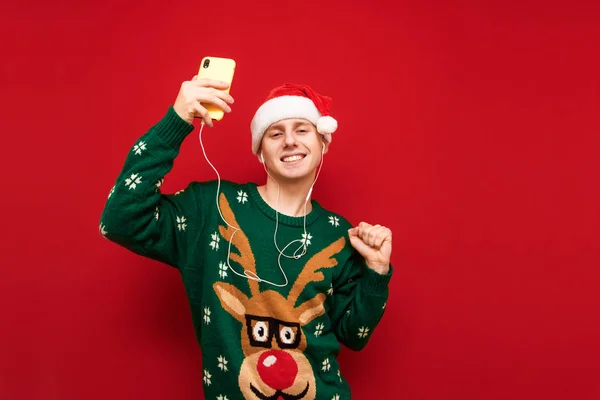 Χαμογελώντας νεαρός άνδρας με smartphone στο χέρι και ακουστικά που διασκεδάζουν στο κόκκινο φόντο, κοιτάζοντας στην κάμερα και χαμογελώντας, φορώντας καπέλο santa και πράσινο. Χριστουγεννιάτικο πουλόβερ. Χριστουγεννιάτικη ιδέα. — Φωτογραφία Αρχείου
