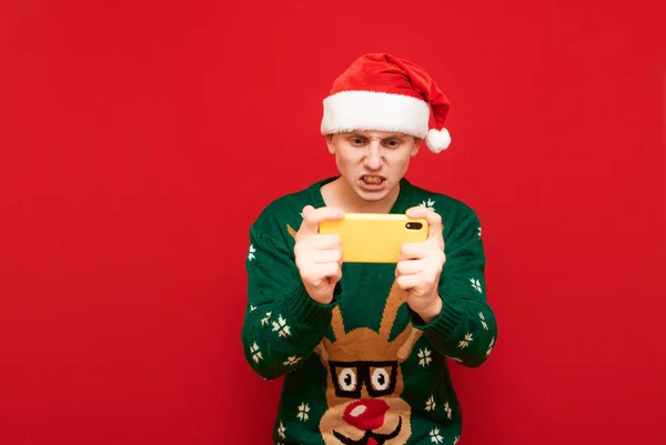 산타 모자를 쓰고 빨간 배경으로 스마트폰으로 게임을 하고 사악 한 얼굴로 스크린을 들여다 보고 따뜻 한 스웨터를 입은 스트레스받는 게이머의 사진. 표현적 인 게임을 하는 비디오 게임. — 스톡 사진
