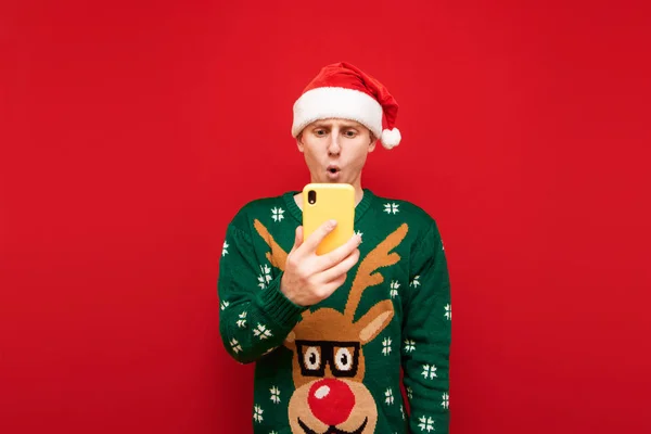 Shocked guy in santa hat and warm green sweater stands on red background and with shocked face looks into screen of smarphone in hand. Surprised young man uses smartphone at Christmas. Isolated.