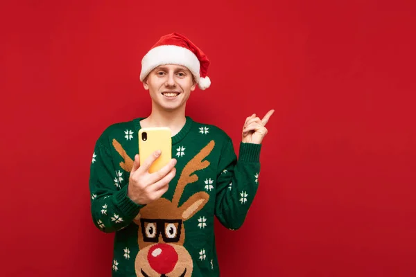웃고 있는 한 젊은 남자가 빨간 배경에 스마트폰을 들고 서서 카메라를 들여다보고 웃으며 복제 공간을 손가락으로 가리키며 크리스마스 스웨터와 산타 모자를 입고 있다. 고립됨 — 스톡 사진