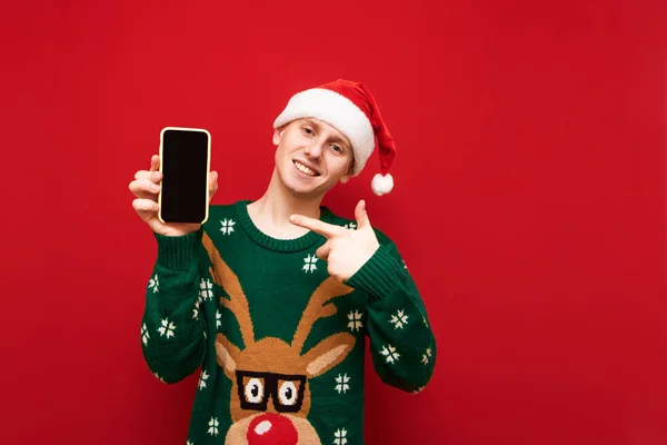 Ευτυχισμένος τύπος με καπέλο santa και χριστουγεννιάτικο πουλόβερ κρατά smartphone στο χέρι του και δείχνει το δάχτυλο στην οθόνη και κοιτάζει στην κάμερα, κόκκινο φόντο. Χαμογελώντας άνθρωπος δείχνει αντίχειρες επάνω στη μαύρη οθόνη smartphone — Φωτογραφία Αρχείου