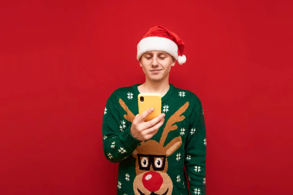 Positive guy in christmas sweater and santa hat uses smartphone on red background, looks at screen and smiles. Christmas boy is using his smartphone. Isolated.