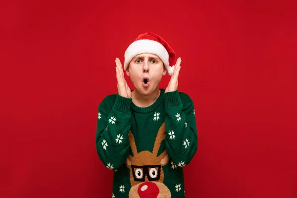 Porträtt av överraskad ung man tittar in i kameran, skriker av chock med upphöjda händer, isolerad på röd bakgrund, bär julen hatt och tröja. Kopiera utrymme — Stockfoto